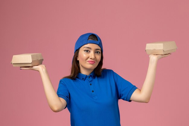 Mensajero femenino de vista frontal con capa uniforme azul sosteniendo pequeños paquetes de entrega en la pared rosa, niña de trabajo de entrega de servicio de empleado