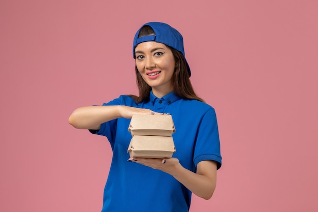 Mensajero femenino de vista frontal en capa uniforme azul sosteniendo pequeños paquetes de entrega en la pared rosa, prestación de servicios para empleados