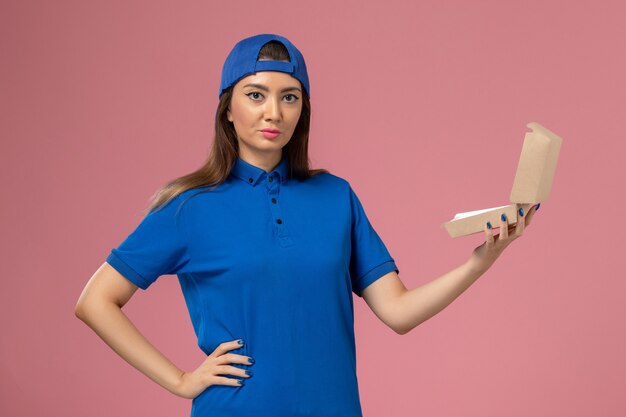 Mensajero femenino de vista frontal en capa uniforme azul sosteniendo pequeños paquetes de entrega en la pared rosa, trabajador de entrega de la empresa de servicios para empleados