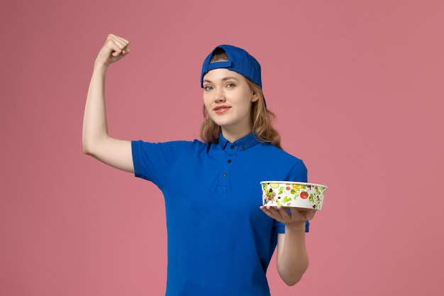 Foto gratuita mensajero femenino de vista frontal en capa uniforme azul sosteniendo el tazón de entrega flexionando en la pared rosa claro, empleado de entrega de servicio