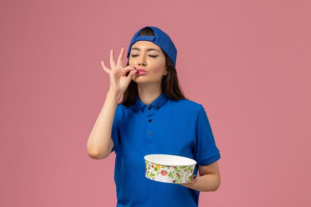 Mensajero femenino de vista frontal en capa uniforme azul sosteniendo el tazón de entrega en la pared rosa claro, entrega de empleados de servicio de trabajo