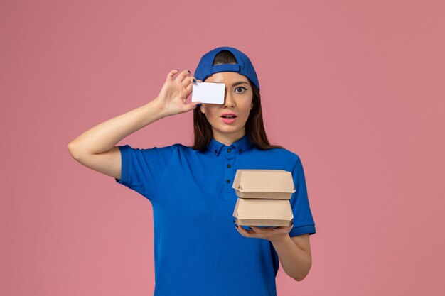 Mensajero femenino de vista frontal en capa uniforme azul con tarjeta y pequeños paquetes de entrega en pared rosa claro, trabajador de entrega de empleados de servicio