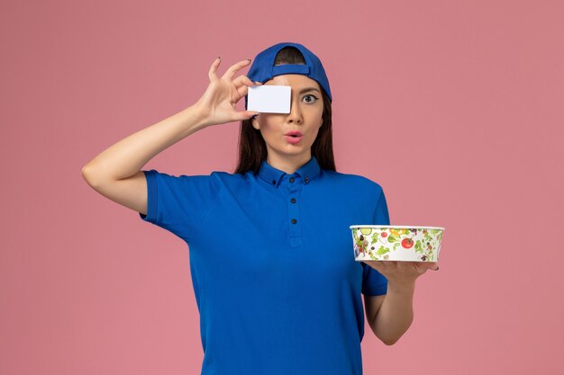 Mensajero femenino de vista frontal en capa uniforme azul con tazón de entrega con tarjeta en la pared de color rosa claro, trabajo de servicio, entrega de empleados