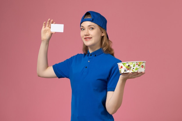 Mensajero femenino de vista frontal en capa uniforme azul con tazón de entrega con tarjeta en la pared rosa claro, empleado de entrega de trabajo de servicio
