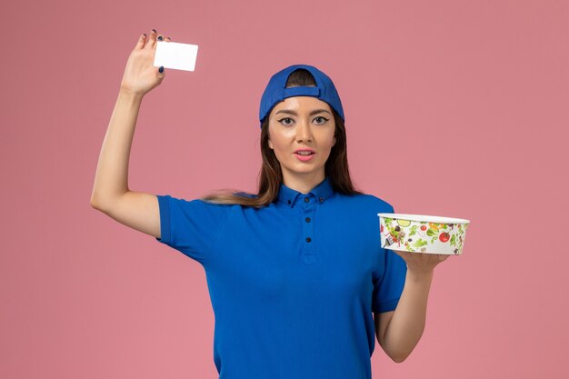 Mensajero femenino de vista frontal en capa uniforme azul con tazón de entrega con tarjeta en la pared rosa claro, entrega de empleados de servicio de trabajo