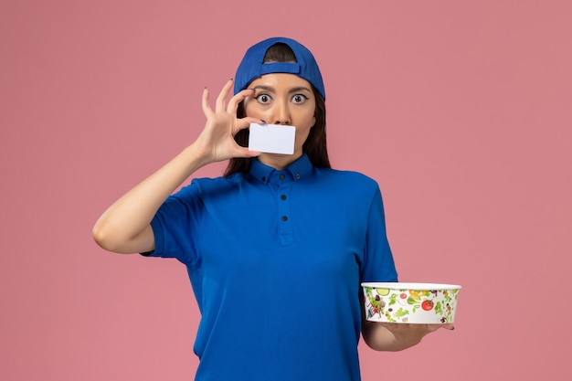 Mensajero femenino de vista frontal en capa uniforme azul con tazón de entrega con tarjeta en la pared rosa claro, entrega de trabajo de trabajo de empleado de servicio