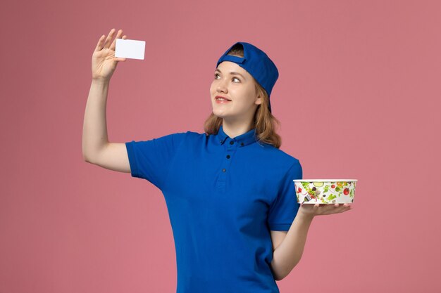 Mensajero femenino de vista frontal en capa uniforme azul con tazón de entrega con tarjeta en la pared rosa claro, trabajador empleado de entrega de servicios