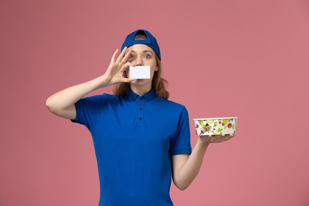 Mensajero femenino de vista frontal en capa uniforme azul con tazón de entrega con tarjeta en la pared rosa claro, trabajo de trabajo de empleado de entrega de servicio