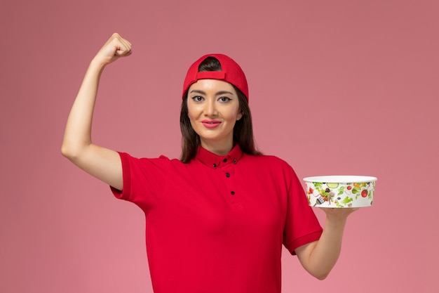 Mensajero femenino de vista frontal en capa uniforme roja con cuenco de entrega en sus manos y flexionando en la pared rosa claro, empleado de entrega uniforme