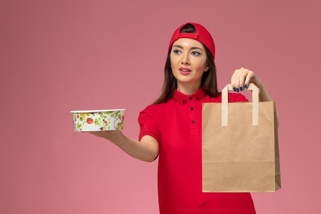 Mensajero femenino de vista frontal en capa uniforme roja con paquete de entrega de papel y cuenco en sus manos en la pared rosa, empleado de trabajo de entrega uniforme