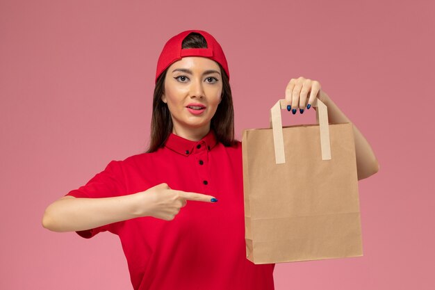 Mensajero femenino de vista frontal en capa uniforme roja con paquete de entrega de papel en sus manos en la pared rosa claro, empleado de trabajo de entrega uniforme