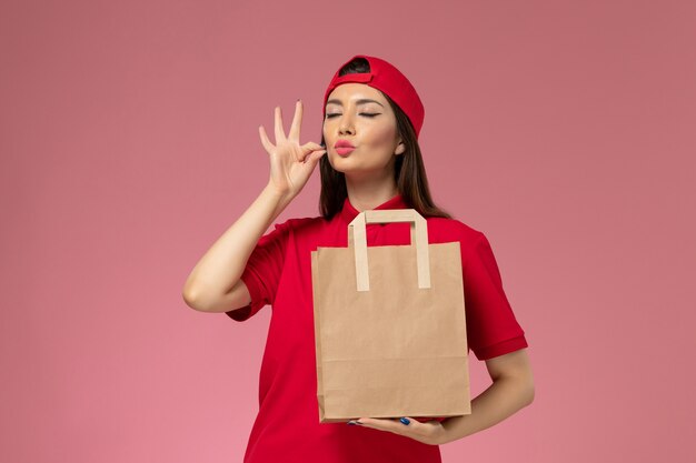 Mensajero femenino de vista frontal en capa uniforme roja con paquete de entrega de papel en sus manos en la pared rosa claro, trabajo de trabajo de empleado de entrega uniforme