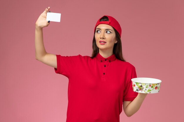 Mensajero femenino de vista frontal en capa uniforme roja con tazón de entrega y tarjeta blanca en sus manos en la pared rosa claro, trabajador empleado de entrega uniforme