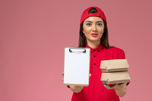Mensajero femenino de vista frontal con capa de uniforme rojo con bloc de notas y pequeños paquetes de comida de entrega en sus manos en la pared rosa claro, trabajo de empleado de entrega de servicio