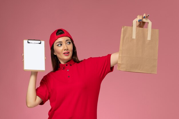 Mensajero femenino de vista frontal en capa de uniforme rojo con paquete de entrega de papel y bloc de notas en sus manos en la pared rosa claro, empleado de entrega uniforme de trabajo