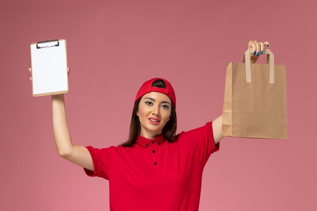 Mensajero femenino de vista frontal en capa de uniforme rojo con paquete de entrega de papel y bloc de notas en sus manos en la pared rosa, empleado de entrega uniforme de trabajo