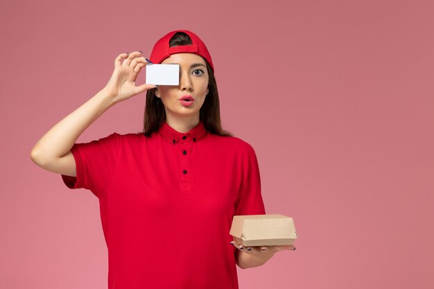 Mensajero femenino de vista frontal con capa de uniforme rojo con pequeño paquete de comida de entrega y tarjeta en sus manos en la pared rosa claro, trabajo de empleado de entrega de servicio