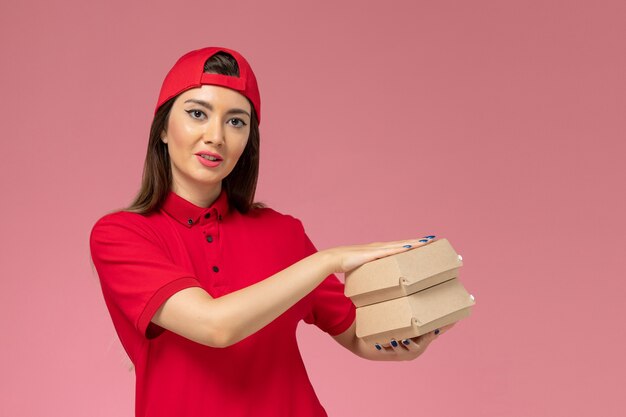 Mensajero femenino de vista frontal con capa de uniforme rojo con pequeños paquetes de comida de entrega en sus manos en la pared rosa claro, trabajo de empleado de entrega de servicio