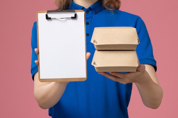 Mensajero femenino de vista frontal cercana en capa uniforme azul sosteniendo pequeños paquetes de comida de entrega y bloc de notas en el empleado de trabajo de servicio de entrega de escritorio rosa