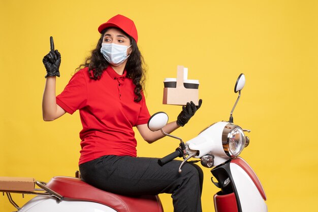 Mensajero femenino de vista frontal en máscara con tazas de café en el fondo amarillo servicio covid- trabajador uniforme de entrega de trabajo