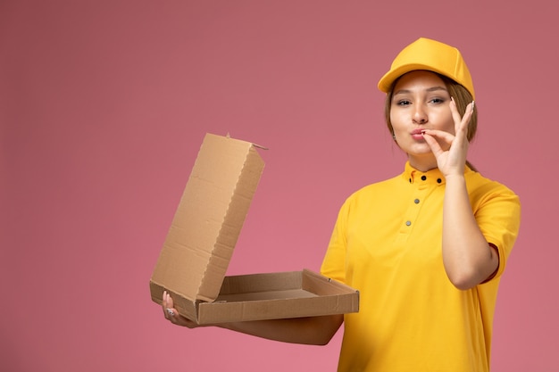 Mensajero femenino de la vista frontal en el paquete de entrega de apertura de capa amarilla uniforme amarillo en el trabajo de trabajo de entrega uniforme de fondo rosa