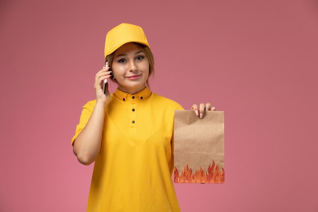 Mensajero femenino de vista frontal en uniforme amarillo capa amarilla sosteniendo el paquete de alimentos blanco hablando por teléfono en el trabajo de color de trabajo de entrega uniforme de escritorio rosa