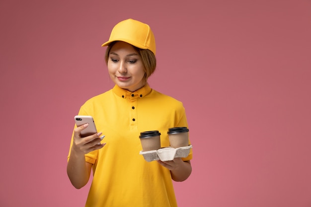 Mensajero femenino de vista frontal en uniforme amarillo capa amarilla sosteniendo tazas de café de plástico usando un teléfono en el color de trabajo de trabajo de entrega uniforme de fondo rosa
