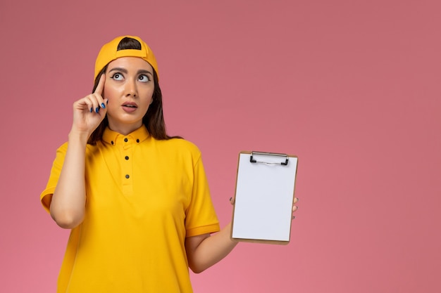 Mensajero femenino de vista frontal en uniforme amarillo y capa con bloc de notas en la entrega uniforme del servicio de empresa de pared rosa