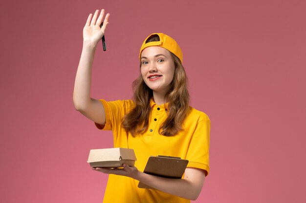 Mensajero femenino de vista frontal con uniforme amarillo y capa con bolígrafo de paquete de comida de entrega y bloc de notas en el uniforme de entrega de servicio de pared rosa claro
