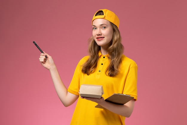 Mensajero femenino de vista frontal con uniforme amarillo y capa con bolígrafo de paquete de comida de entrega y bloc de notas en el uniforme de entrega de servicio de pared rosa claro