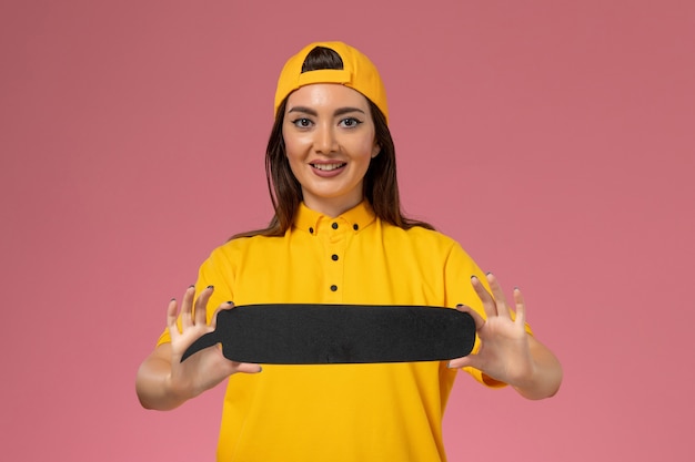 Mensajero femenino de vista frontal en uniforme amarillo y capa con cartel negro en la chica de entrega uniforme de servicio de empresa de pared rosa