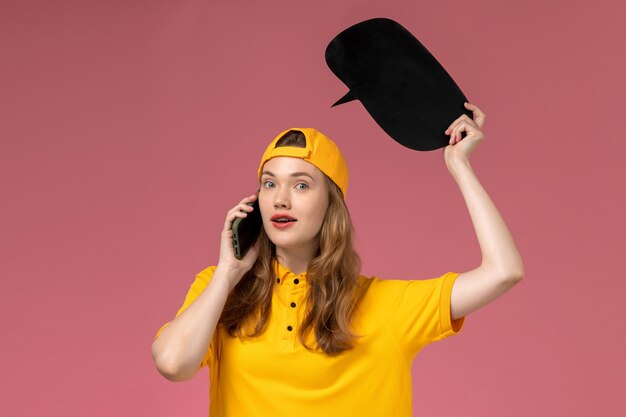 Foto gratuita mensajero femenino de vista frontal en uniforme amarillo y capa con cartel negro y hablando por teléfono en el uniforme de entrega de trabajador de servicio de empresa de pared rosa