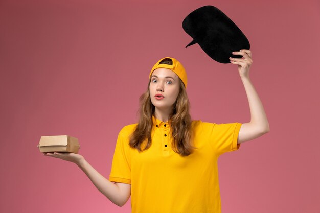 Mensajero femenino de vista frontal en uniforme amarillo y capa con cartel negro con paquete de alimentos en uniforme de entrega de trabajador de servicio de empresa de pared rosa