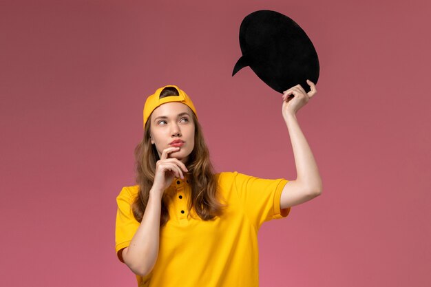 Mensajero femenino de vista frontal en uniforme amarillo y capa con cartel negro pensando en trabajo de empresa uniforme de entrega de servicio de pared rosa claro