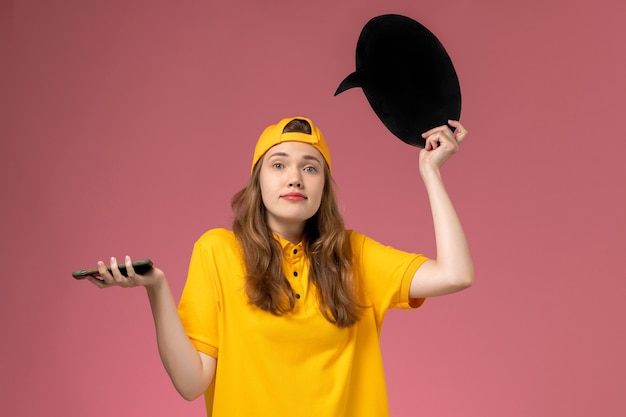 Mensajero femenino de vista frontal en uniforme amarillo y capa con cartel negro y teléfono inteligente en uniforme de trabajo de entrega de servicio de empresa de pared rosa