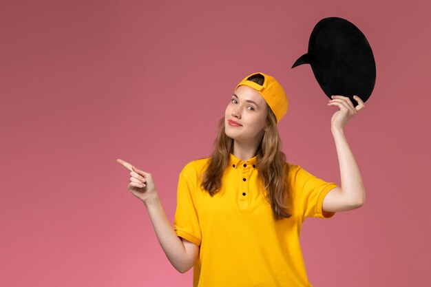 Mensajero femenino de vista frontal en uniforme amarillo y capa con cartel negro en el trabajo uniforme de entrega de servicio de empresa de pared rosa