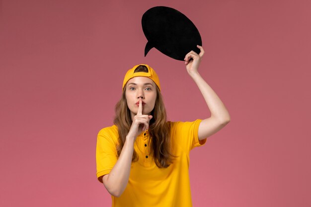 Mensajero femenino de vista frontal en uniforme amarillo y capa con gran cartel negro en la pared rosa, servicio de empresa trabajo uniforme de entrega