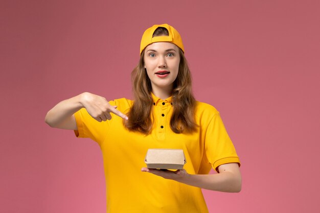 Mensajero femenino de vista frontal en uniforme amarillo y capa con paquete de comida de entrega en la pared rosa servicio de entrega uniforme trabajador de la empresa