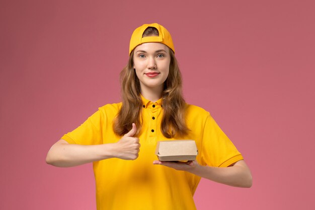 Mensajero femenino de vista frontal en uniforme amarillo y capa con paquete de comida de entrega en el trabajo de empresa uniforme de entrega de servicio de pared rosa