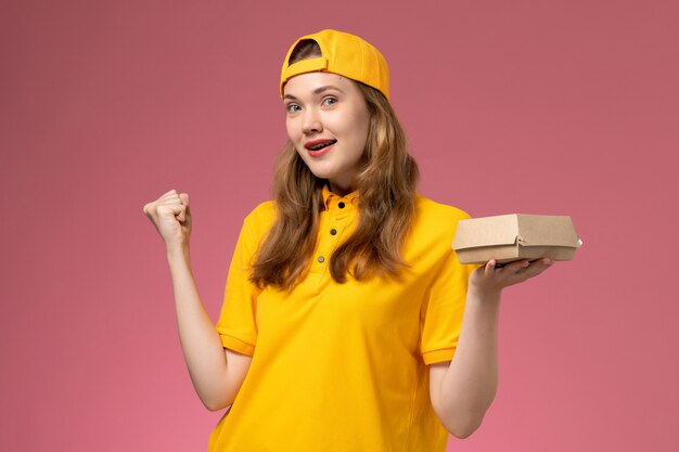 Mensajero femenino de vista frontal en uniforme amarillo y capa con paquete de comida de entrega en el trabajo uniforme de entrega de servicio de pared rosa