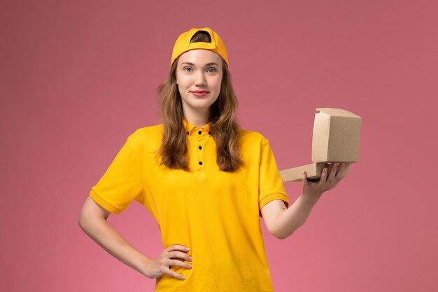 Mensajero femenino de vista frontal en uniforme amarillo y capa con pequeña apertura de paquete de comida de entrega en uniforme de entrega de trabajo de trabajo de servicio de pared rosa claro