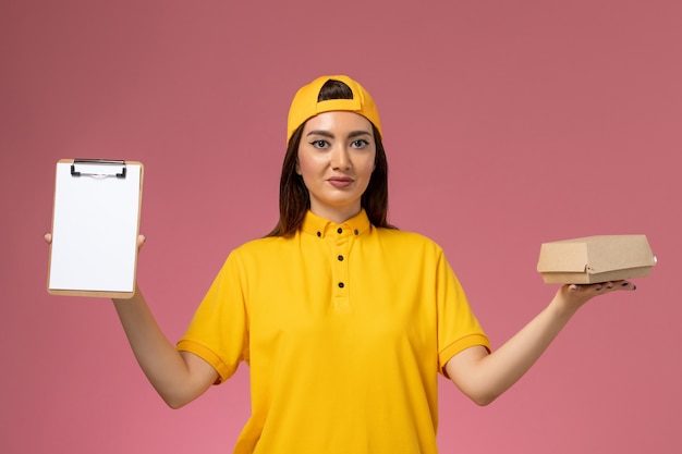 Mensajero femenino de vista frontal en uniforme amarillo y capa con pequeño paquete de comida de entrega y bloc de notas en el trabajo de trabajo de entrega uniforme de servicio de pared rosa claro