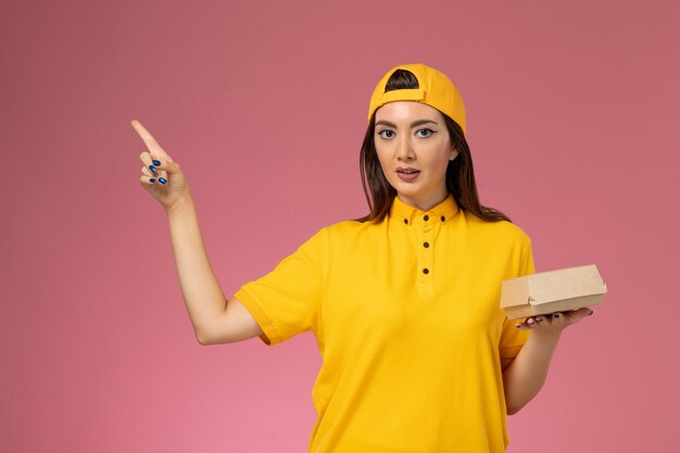 Mensajero femenino de vista frontal en uniforme amarillo y capa con pequeño paquete de comida de entrega en la empresa de entrega de servicio de niña uniforme de pared rosa