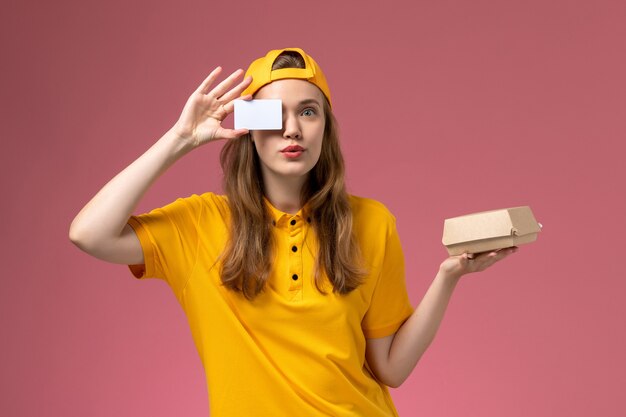 Mensajero femenino de vista frontal con uniforme amarillo y capa con pequeño paquete de comida de entrega y tarjeta de plástico en uniforme de entrega de servicio de trabajo de pared rosa