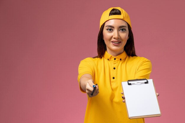 Mensajero femenino de la vista frontal en uniforme amarillo y capa que sostiene el bloc de notas y el bolígrafo en el trabajo de entrega uniforme de servicio de empresa de pared rosa claro