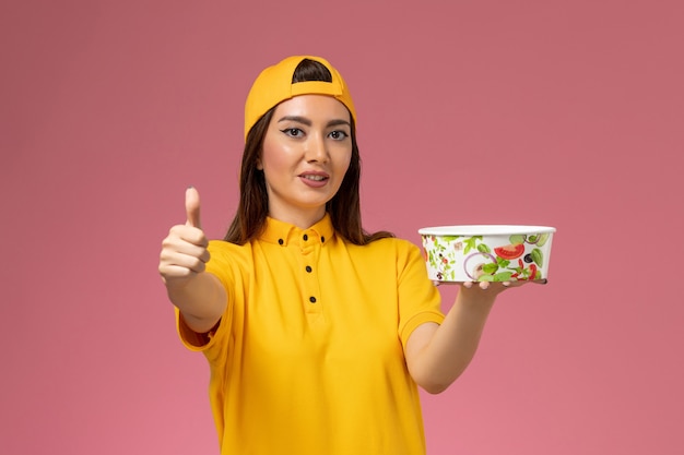 Mensajero femenino de la vista frontal en uniforme amarillo y capa que sostiene el cuenco de entrega redondo en la entrega uniforme del servicio de la pared rosada