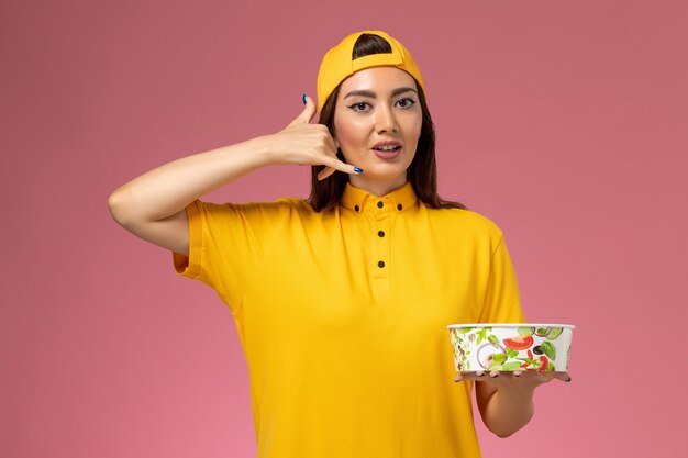 Mensajero femenino de vista frontal en uniforme amarillo y capa que sostiene el cuenco de entrega redondo en la entrega uniforme del servicio de trabajo de la pared rosada