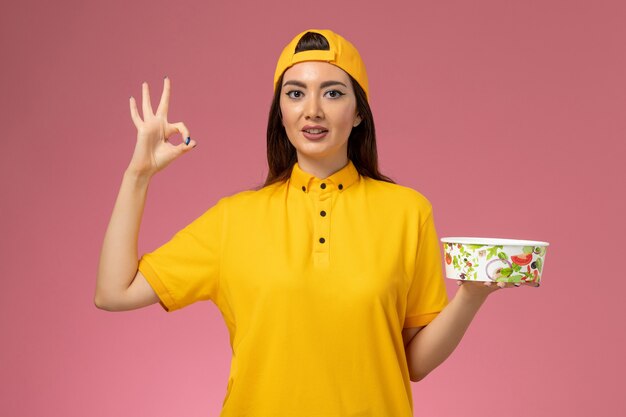 Mensajero femenino de la vista frontal en uniforme amarillo y capa que sostiene el cuenco de entrega redondo en la muchacha del trabajador de entrega uniforme del servicio de la pared rosada