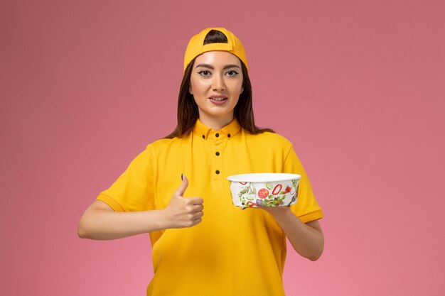 Mensajero femenino de vista frontal en uniforme amarillo y capa que sostiene el cuenco de entrega redondo en el trabajo de trabajo de entrega uniforme de servicio de pared rosa