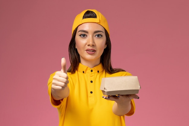 Mensajero femenino de la vista frontal en uniforme amarillo y capa que sostiene el pequeño paquete de comida de entrega en la empresa de niña de trabajador de entrega de servicio uniforme de pared rosa